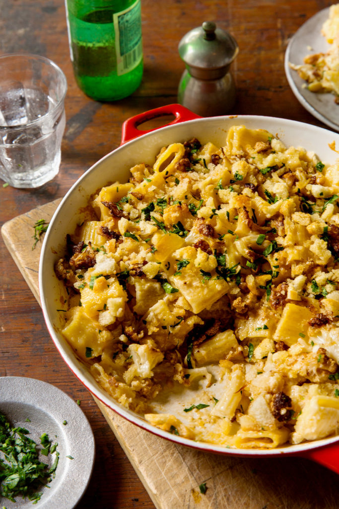 Pan full of cauliflower baked ziti surrounded by a dish of herbs, glass of water, bottle of sparkling water, and pepper grinder. 